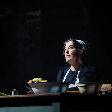 A woman dressed as a housekeeper looks out to the audience, she sits at a table, with vegetables on display.