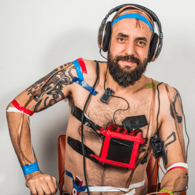 A man sitting on a chair covered in wires and tape, wearing a pair of headphones.