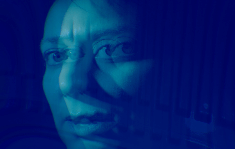 A woman’s face looks directly to camera, over a vivid blue background. A mirror image overlaps her face creating a distorted effect.