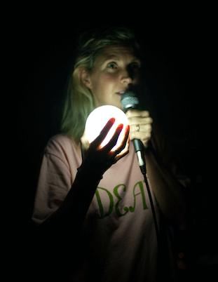 A woman holding a globe and microphone in the dark, the globe lights up her face.