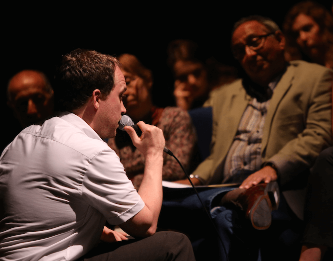 A man speaking into a microphone to an audience who can be seen in their seats.