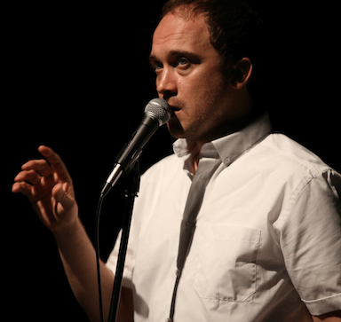 A man speaking into a microphone with his right hand raised slightly.