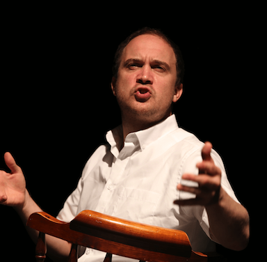 A man with his hands raised speaking and looking up towards a spot light.