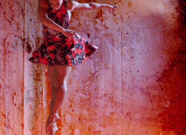 A woman jumping and twisting her body in a dance pose, against a ink splattered projection.