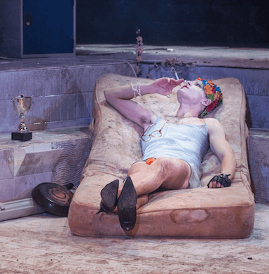 A person lying on a single mattress, in an empty swimming pool, smoking, wearing heels and a flowery swimming cap.