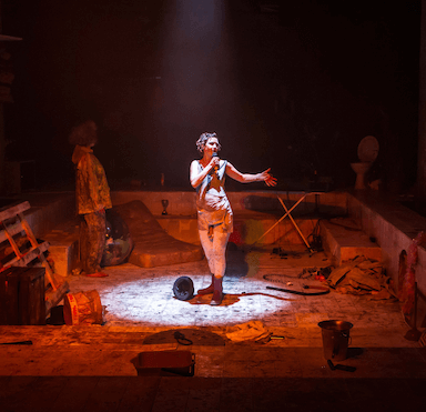 A woman standing in an empty swimming pool, varies items lie around her, she is lit up by a circular spot light with a microphone to her mouth.