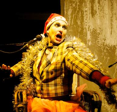 A person dressed in a chequered jacket, wearing a Christmas hat and tinsel over his shoulders, his head is turned away from a microphone.