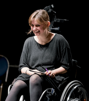 A woman sitting in a wheelchair, not looking at the camera, with a pen and paper in hand.