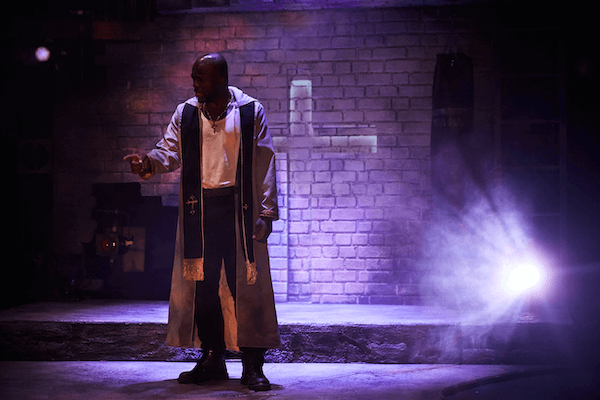 A young man dressed in a Friar outfit with his hand raised. There is a cross visible on a brick wall behind him.