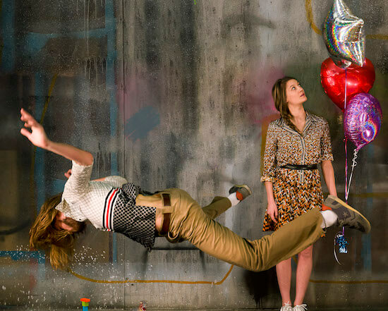 A man levitating in the air face down, a woman stands behind him holding three helium balloons.