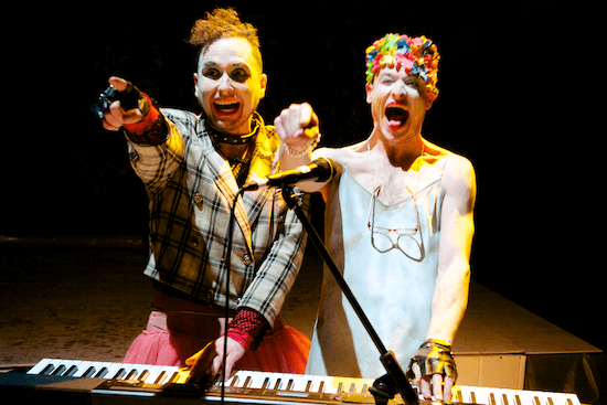 Two people standing at a keyboard, laughing and pointing out towards the camera.