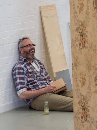 A man sitting on the floor, leaning against a white brick wall, holding a notebook. He appears to be laughing at something out of view of the camera.
