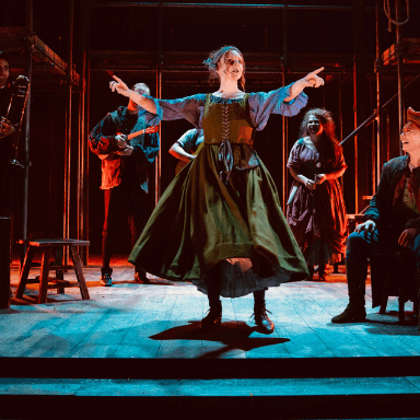 A woman standing on stage point her arms and hands out towards the audience.