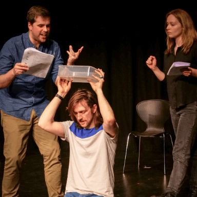 Two people holding scripts gesture towards a man holding a plastic tub above his head kneeling on the floor.