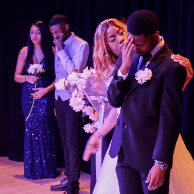 Two people stand in the background, whilst a bride stands looking at her groom who has his head down.