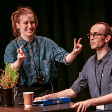 A man sits holding the edge of a table, as he looks out to the audience with concern, whilst a woman stands next to him doing inverted commas with her hands.