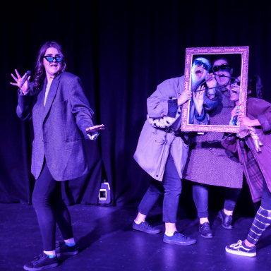 A group of people wearing jackets and sunglasses, holding poses through a frame, one stands off to the left.