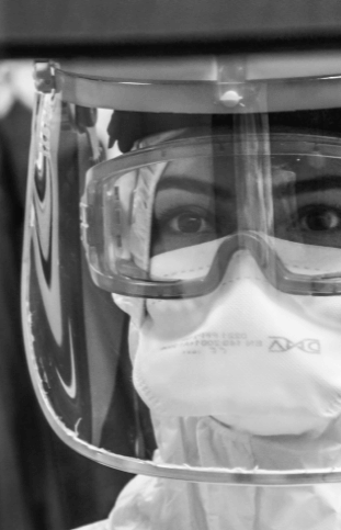 A black and white photograph of a persons face behind a plastic shield and glasses, dressed in full PPE.