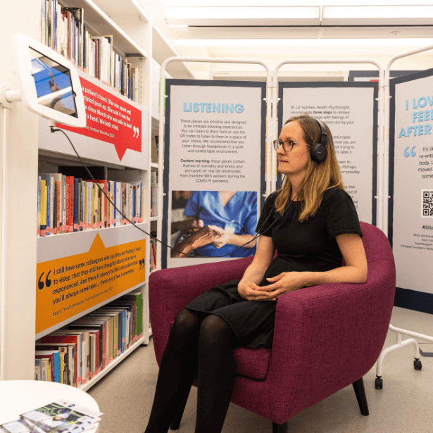 A woman wearing headphones sits in an armchair watching a small screen attached to the wall. Behind her are shelves of books.