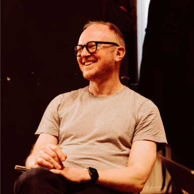 A man wearing black rimmed glasses, sitting with his hands on his knee. He appears to be smiling at someone out of shot.