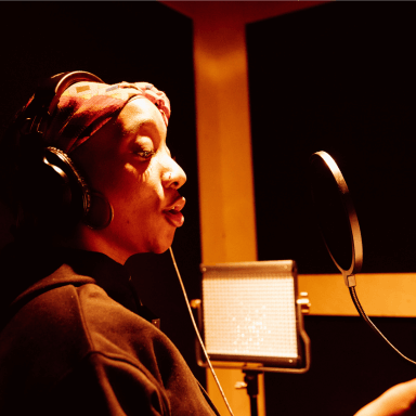 A young woman wearing headphones, singing into a microphone in a studio booth.
