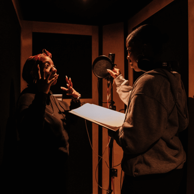 Two young woman stand in a studio booth, facing one another.
