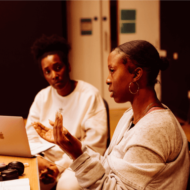 A side profile of a young black woman, who is talking to people out of shot. In the background another person is looking towards the woman listening.