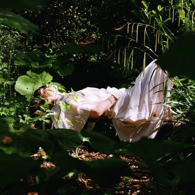 A person in a white dress lies on the ground surrounded by plants.