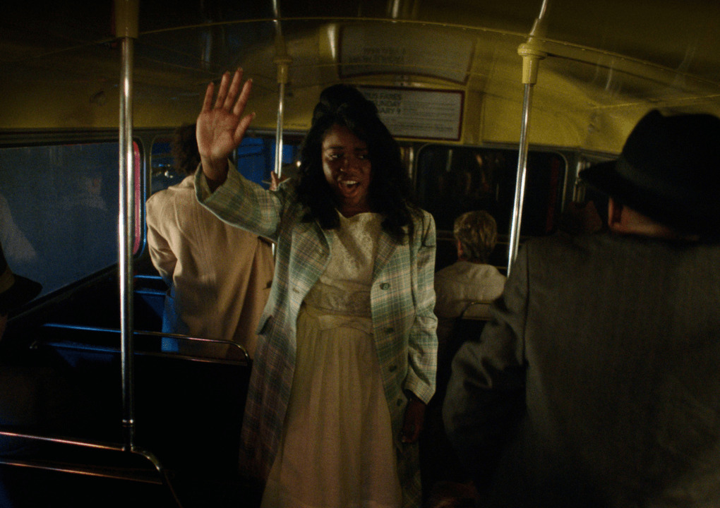 A woman standing on a bus holding her right hand in the air whilst singing.