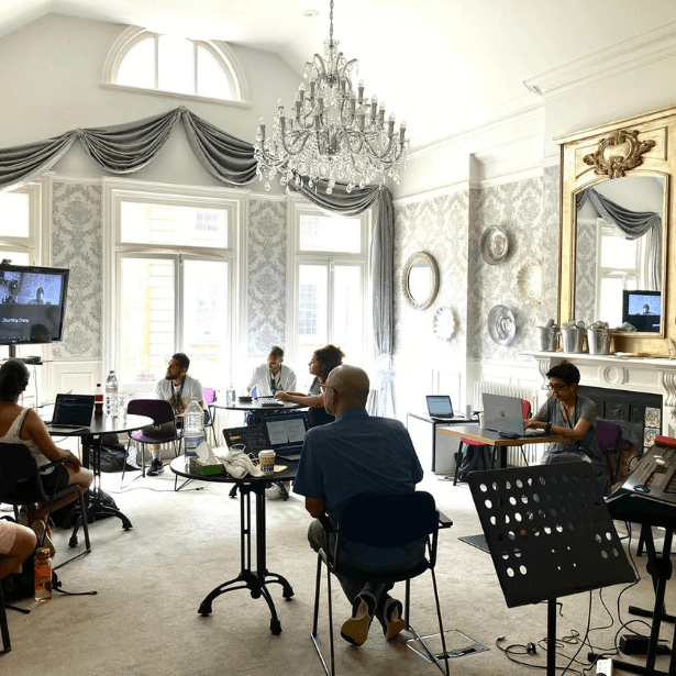 A group of people sitting around laptops watching and listening to people on a TV screen.