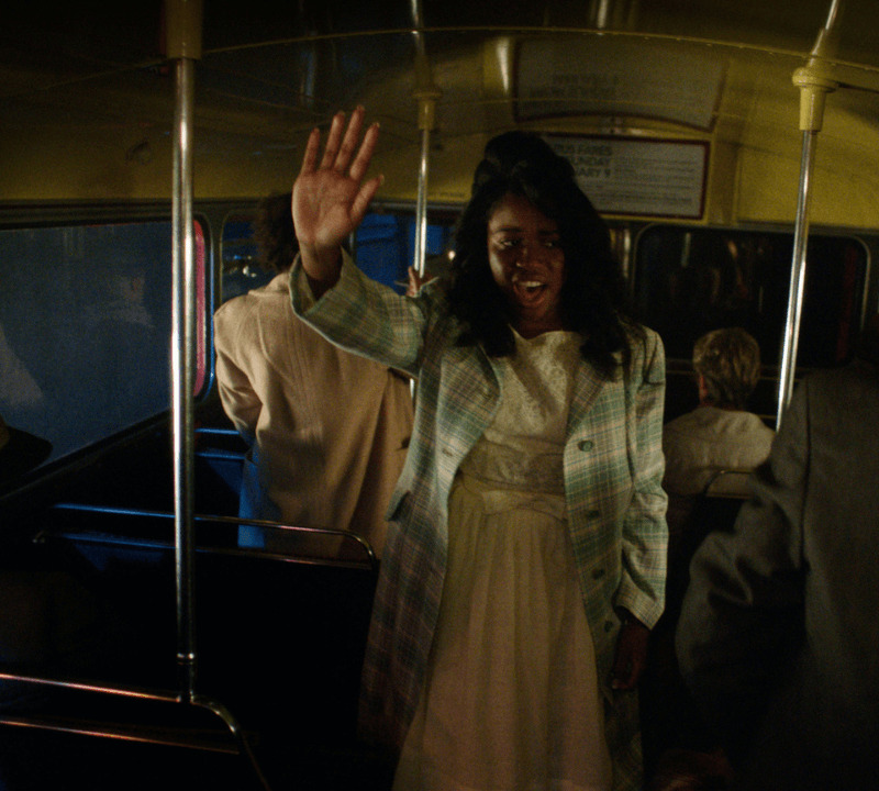 A woman standing on a bus holding her right hand in the air whilst singing.