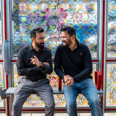 Two people are sat on the seats in front of a bus stop shelter decorated with stain glass style patterns.
