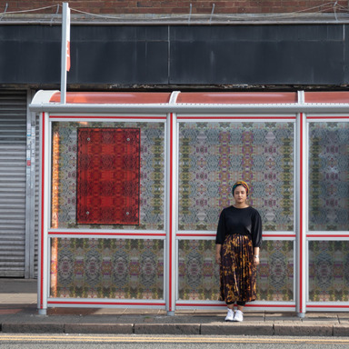 Photograph of a bus stop shelter in front of a shop front, decorated with stain glass style patterns. A person stands in front of it facing the camera.