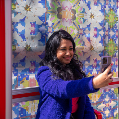 A person sits in front of a bus stop shelter decorated with stain glass style patterns, taking a selfie