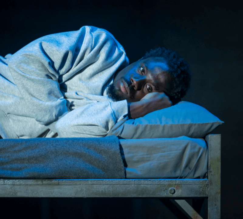 A young black man wearing a grey tracksuit is lay down on a bed. He has his arm tucked under his head.