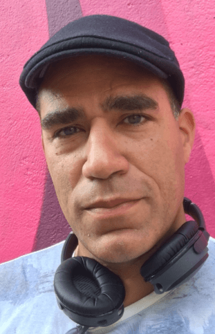 A photograph of a man wearing a flat cap and headphones round his neck, looking directly to the camera.