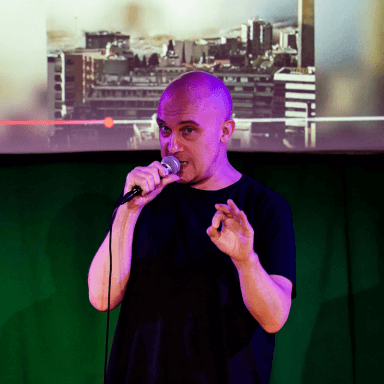 A photograph of Chris Thorpe stood holding a microphone to his mouth. Behind him is a partial view of a projection screen.