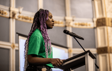 A person with long braided purple hair, wearing a bright green top, stands at a lectern speaking into a microphone.