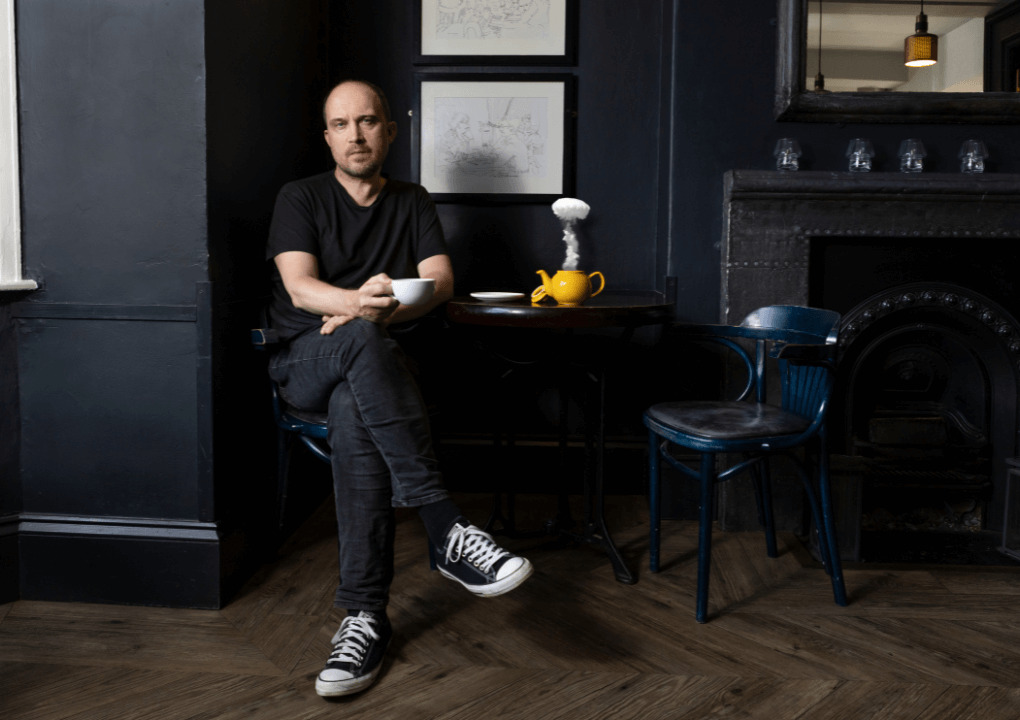 A man sitting at a coffee table, holding a mug, with a yellow teapot on the table with smoke escaping.