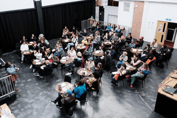 An aerial view of a group of people sat round small circular tables, facing a panel of five people sat side by side on a raised platform.