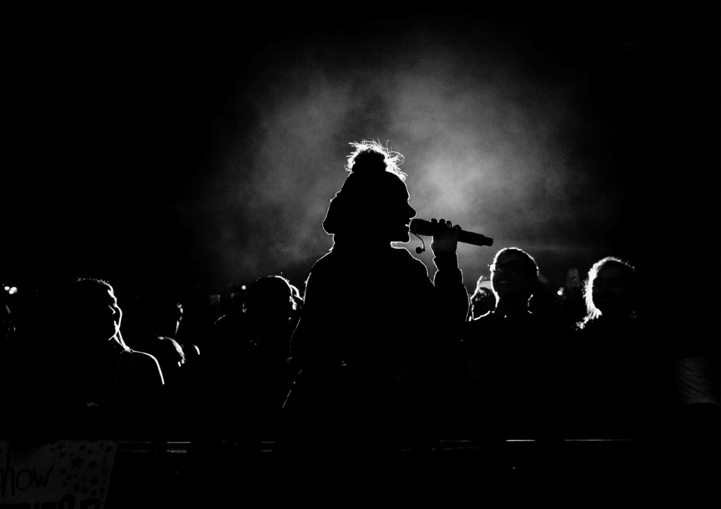A side profile of a person singing with a microphone to their mouth. The bright light behind them has created a silhouette of them and the audience.