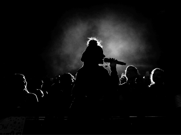 A side profile of a person singing with a microphone to their mouth. The bright light behind them has created a silhouette of them and the audience.