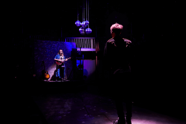A young woman stands in a spotlight, singing into a microphone, playing a guitar. She is facing a young man who has his back to the camera.