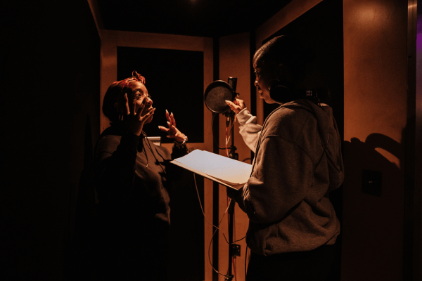 Two young woman stand in a studio booth, facing one another.