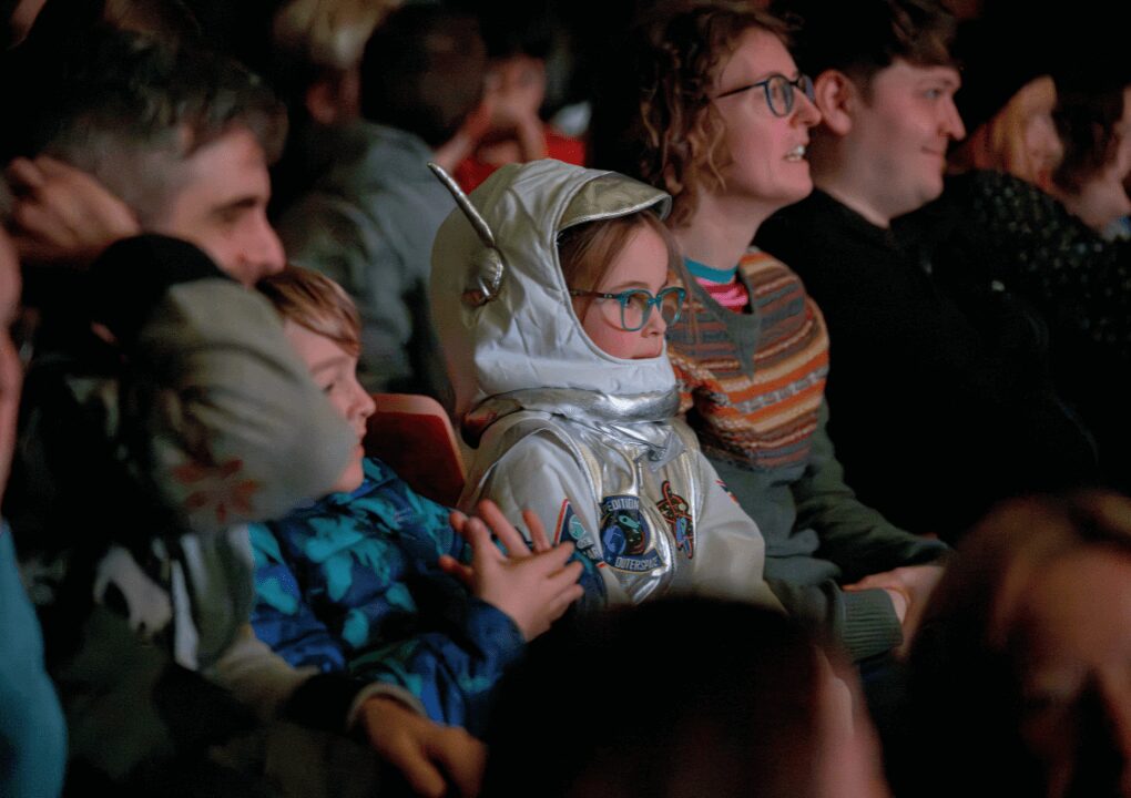 A young girl is sat in the audience, dressed as an astronaut, looking towards the stage out of shot.