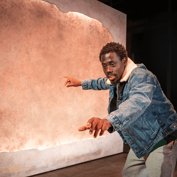A young black man dressed in a denim jacket and grey joggers. He is slightly hunched over pointing at a painting behind him.